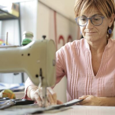 Focused mature female in casual clothes and eyeglasses sitting at table and sewing leather purse while working in modern workshop